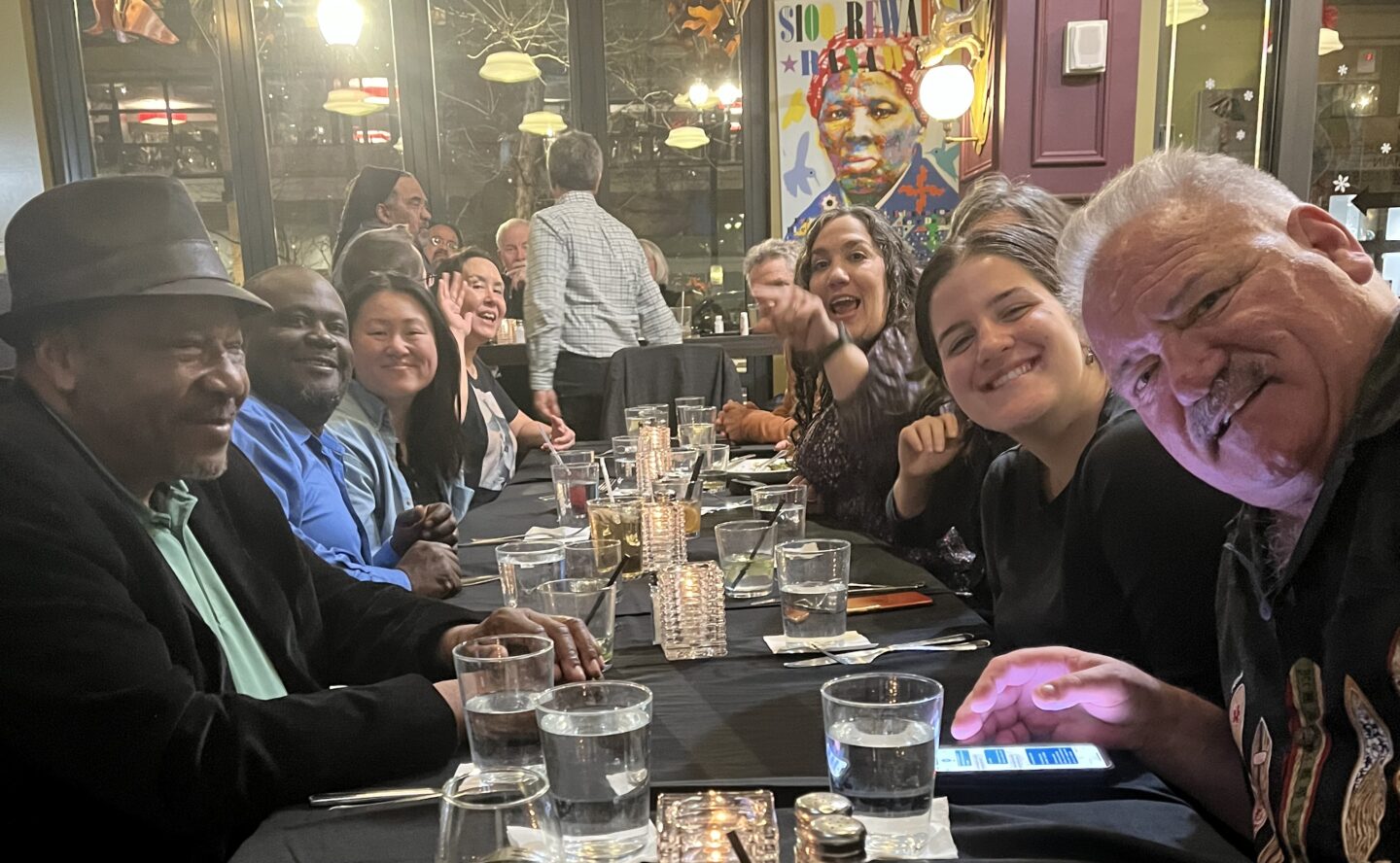 DCO2 members enjoy a lively dinner together at Busboys and Poets in Washington, DC. Everyone is smiling. A colorful painting of Harriet Tubman appears on a wall in the background.