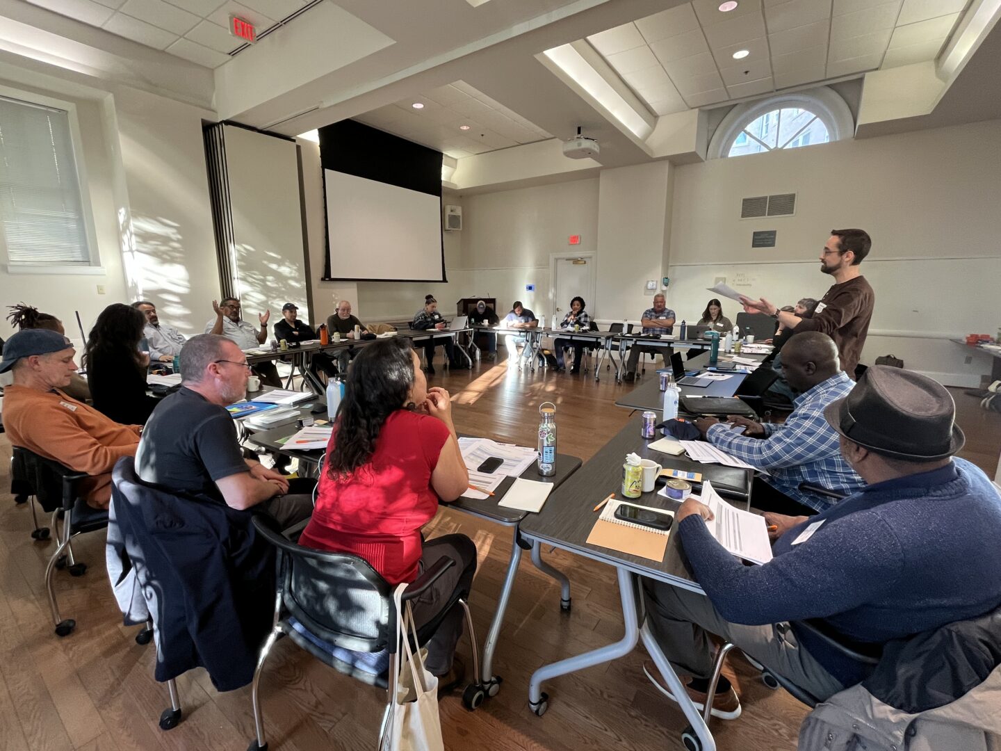 DCO2 Legislative Director James Mitchell facilitates a strategy session during the annual member fly-in. In a large meeting room, desks are arranged in a square configuration, with members seated along the outer edges facing inward. A projection screen is visible in the background. James stands holding a piece of paper, addressing the group.