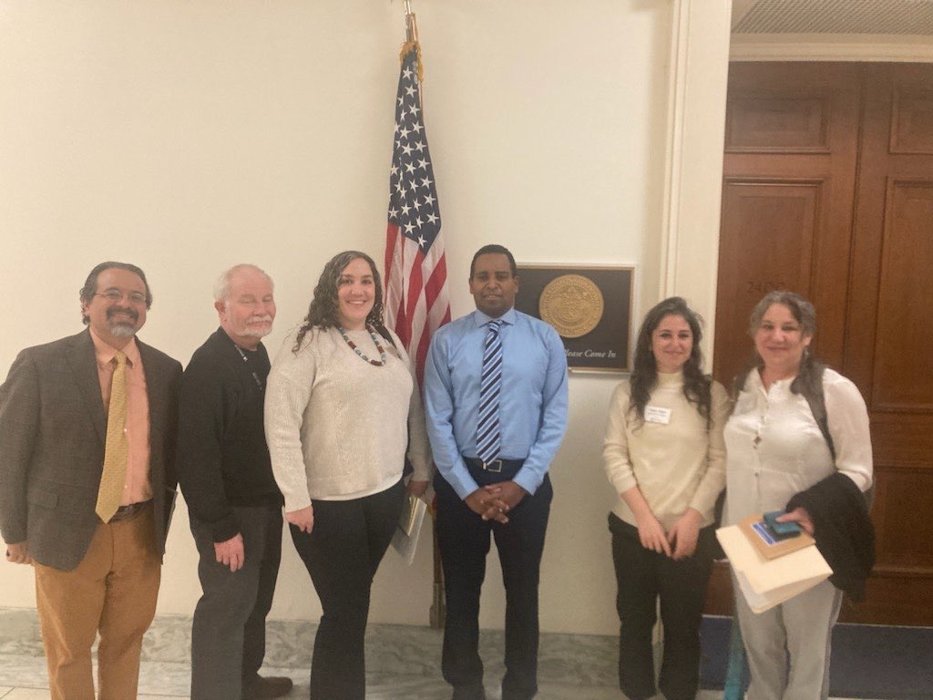 Rep. Joe Neguse stands in the hallway outside of his office in Washington, DC, alongside five members and staff of DCO2. The group is smiling, and an American flag sits on a flagpole behind them.