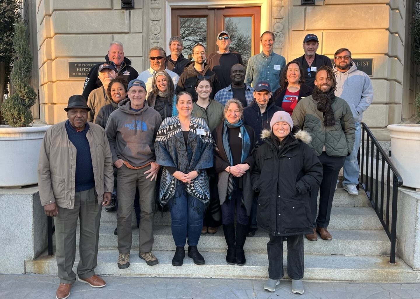 DCO2 members standing together at sunset on the stairs of the United Methodist Building in Washington, DC. Everyone is smiling and wearing winter clothing, including hats, sweatshirts, and coats.